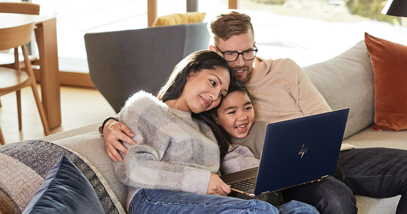 Двое взрослых и ребенок сидят в обнимку на диване, смотрят на что-то на экране ноутбука и улыбаются.